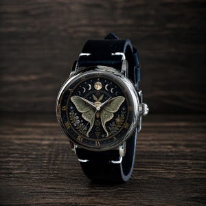 May include: A round wristwatch with a black leather band. The watch face features a detailed illustration of a moth with a celestial design, including phases of the moon and floral accents. The watch has silver-colored metal casing.