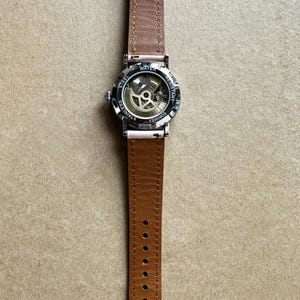 May include: A silver wristwatch with a brown leather band. The watch has a see-through back revealing the inner workings of the watch. The watch face says 'Water Resistant' and 'Stainless Steel'.