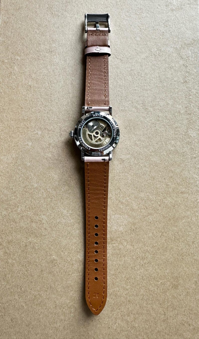May include: A silver wristwatch with a brown leather band. The watch has a see-through back revealing the inner workings of the watch. The watch face says 'Water Resistant' and 'Stainless Steel'.