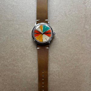 Op de afbeelding: Een ronde polshorloge met een bruine leren band. De wijzerplaat is verdeeld in kleurrijke segmenten van rood, oranje, geel, groen en cr&egrave;me. Het horloge heeft een zilverkleurige kast en een gespsluiting. De band is ca. 22 cm lang.