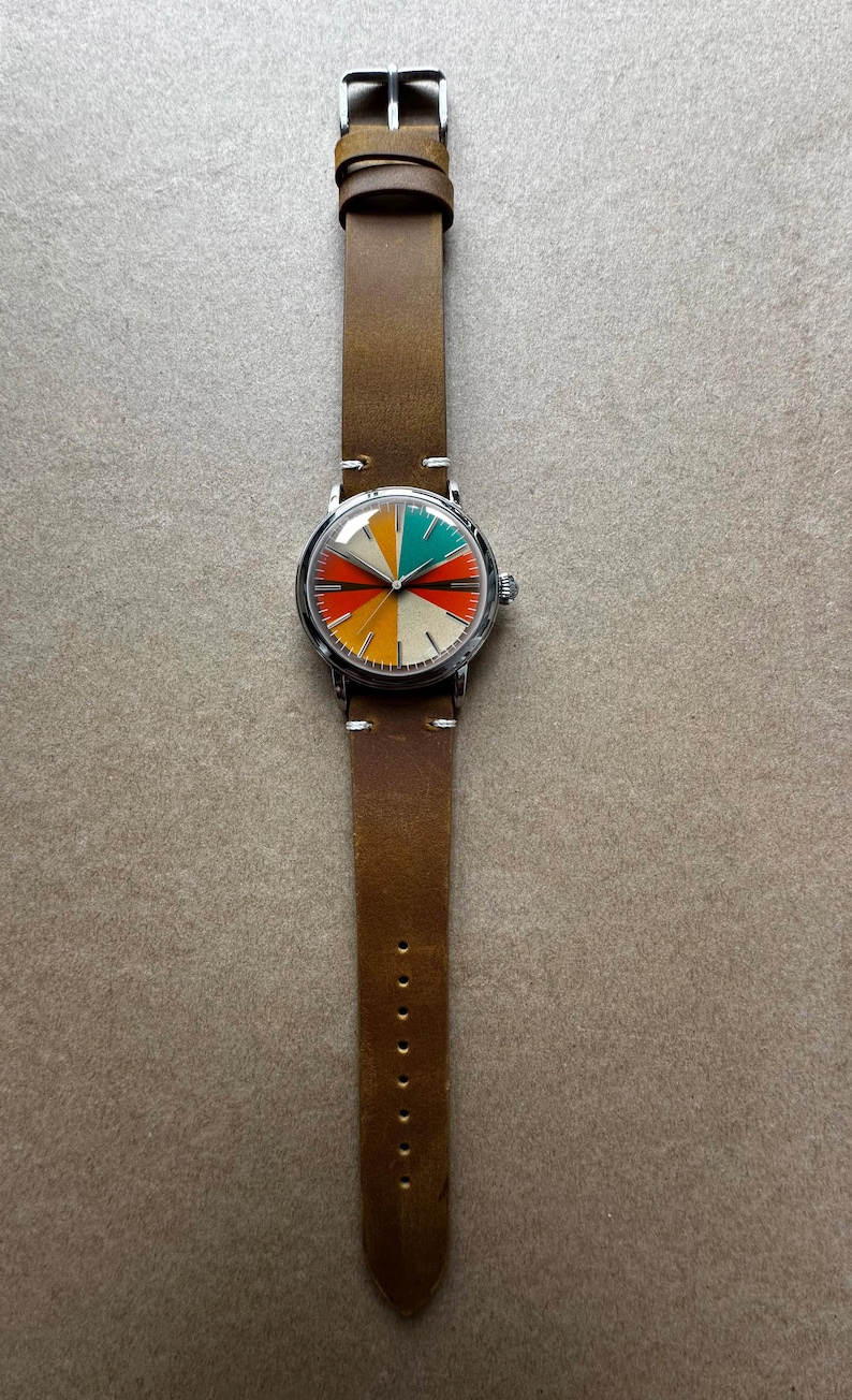 Op de afbeelding: Een ronde polshorloge met een bruine leren band. De wijzerplaat is verdeeld in kleurrijke segmenten van rood, oranje, geel, groen en cr&egrave;me. Het horloge heeft een zilverkleurige kast en een gespsluiting. De band is ca. 22 cm lang.