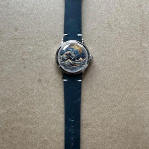 May include: A wristwatch with a blue leather band and a silver-colored case. The watch face features a detailed illustration of a wave scene with a boat. The band is approximately 9 inches long.
