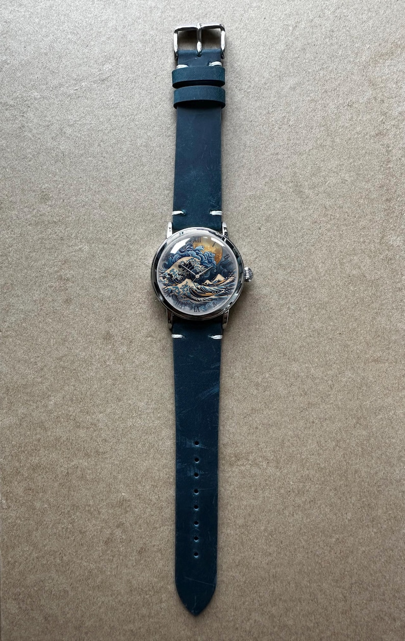May include: A wristwatch with a blue leather band and a silver-colored case. The watch face features a detailed illustration of a wave scene with a boat. The band is approximately 9 inches long.
