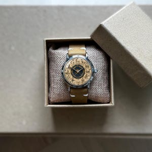 Puede incluir: Un reloj de pulsera con una correa de cuero marr&oacute;n claro y una caja plateada. La esfera del reloj presenta un dise&ntilde;o detallado con figuras de animales. El reloj se presenta en una peque&ntilde;a caja de cart&oacute;n abierta con un coj&iacute;n tipo arpillera.
