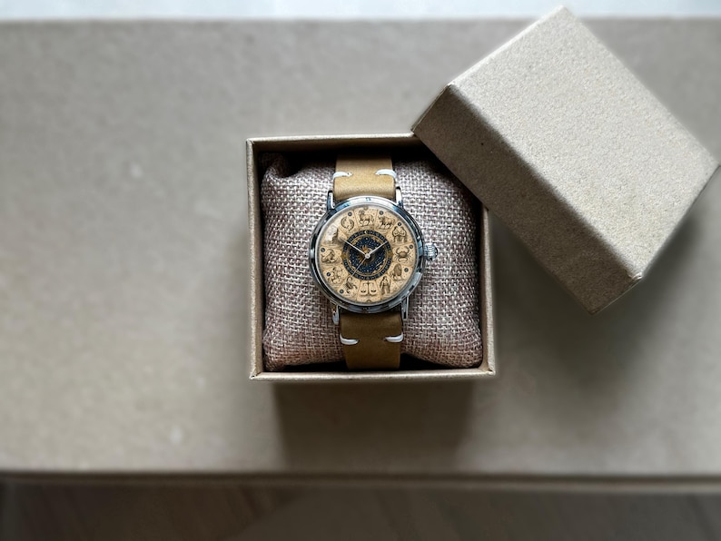 Puede incluir: Un reloj de pulsera con una correa de cuero marr&oacute;n claro y una caja plateada. La esfera del reloj presenta un dise&ntilde;o detallado con figuras de animales. El reloj se presenta en una peque&ntilde;a caja de cart&oacute;n abierta con un coj&iacute;n tipo arpillera.
