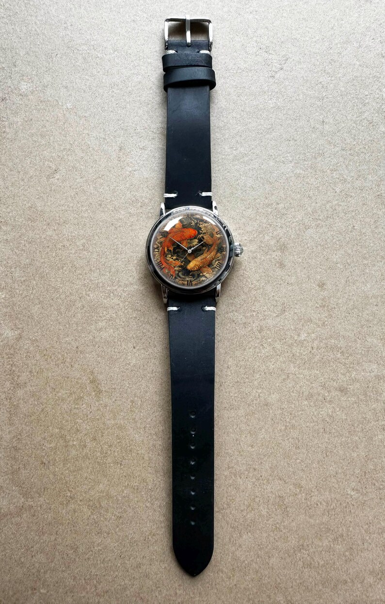 May include: A round wristwatch with a silver case and a black leather band. The watch face features a detailed image of koi fish in shades of orange and gold. The band is approximately 1 inch wide.