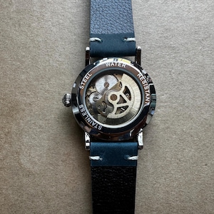 May include: A stainless steel watch with a visible mechanical movement. The watch has a dark blue leather strap and a silver-colored case. The back of the watch is transparent, displaying the intricate gears. The watch is water resistant.