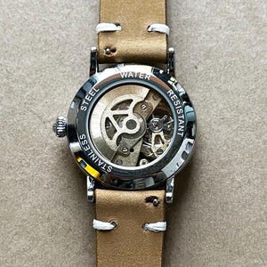 Puede incluir: Primer plano de un reloj de acero inoxidable con una correa de cuero marr&oacute;n claro. La esfera del reloj es transparente, revelando el mecanismo interno. El reloj est&aacute; etiquetado como "Water Resistant" y "Stainless Steel". La correa tiene costuras blancas.