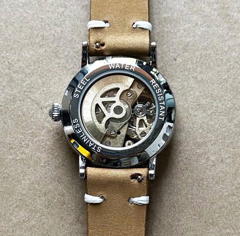 Puede incluir: Primer plano de un reloj de acero inoxidable con una correa de cuero marr&oacute;n claro. La esfera del reloj es transparente, revelando el mecanismo interno. El reloj est&aacute; etiquetado como "Water Resistant" y "Stainless Steel". La correa tiene costuras blancas.