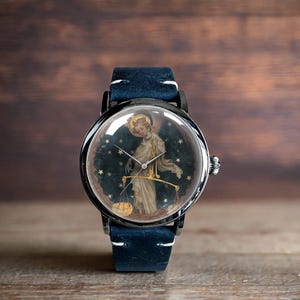 May include: A silver wristwatch with a blue leather band. The watch face features a painting of an angel holding a scale.