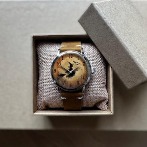 May include: A wristwatch with a brown leather band and a silver-colored case. The watch face features a witch on a broomstick design with a tan background. The watch is presented in a small, open cardboard box.