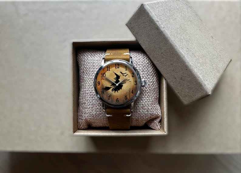May include: A wristwatch with a brown leather band and a silver-colored case. The watch face features a witch on a broomstick design with a tan background. The watch is presented in a small, open cardboard box.