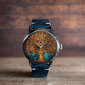 Puede incluir: Un reloj de pulsera redondo con caja negra y correa de cuero azul. La esfera del reloj presenta un dise&ntilde;o detallado de &aacute;rbol en tonos dorados, marrones y azules. El reloj tiene una corona plateada y costuras blancas en la correa.