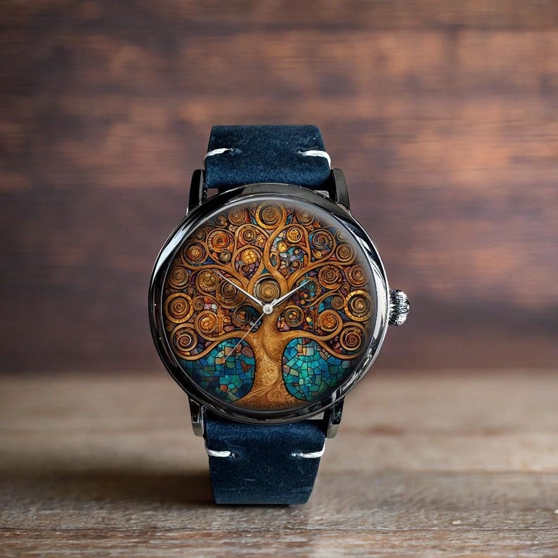 Puede incluir: Un reloj de pulsera redondo con caja negra y correa de cuero azul. La esfera del reloj presenta un dise&ntilde;o detallado de &aacute;rbol en tonos dorados, marrones y azules. El reloj tiene una corona plateada y costuras blancas en la correa.