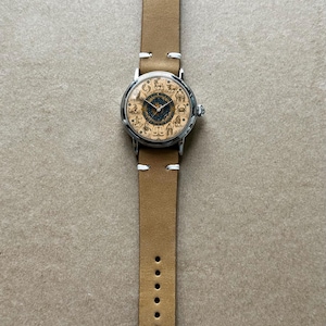 Puede incluir: Reloj de pulsera con correa de cuero marr&oacute;n claro y caja plateada. La esfera del reloj presenta s&iacute;mbolos del zodiaco sobre un fondo beige con un centro azul oscuro. La correa tiene costuras blancas y una hebilla plateada.