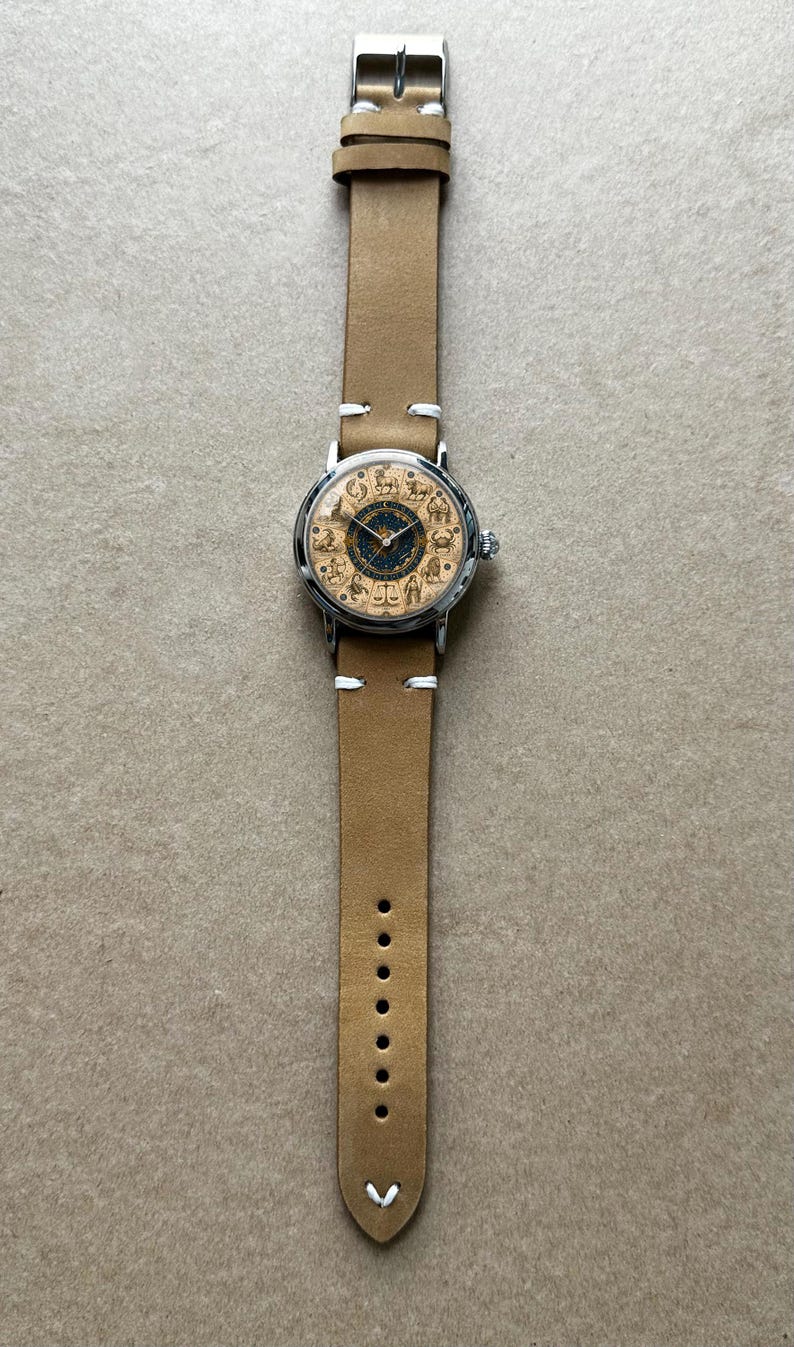 Puede incluir: Reloj de pulsera con correa de cuero marr&oacute;n claro y caja plateada. La esfera del reloj presenta s&iacute;mbolos del zodiaco sobre un fondo beige con un centro azul oscuro. La correa tiene costuras blancas y una hebilla plateada.