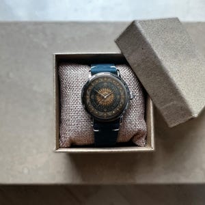 May include: A round wristwatch with a detailed face featuring a sunburst design and a dark blue leather band. The watch is presented in a small, open cardboard box with a soft, beige cushion.