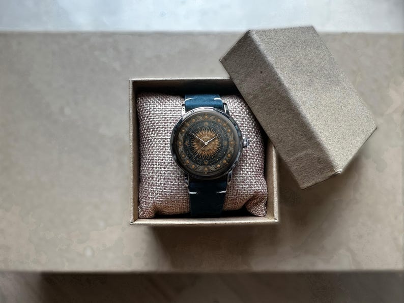 May include: A round wristwatch with a detailed face featuring a sunburst design and a dark blue leather band. The watch is presented in a small, open cardboard box with a soft, beige cushion.