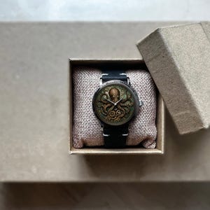 May include: A steampunk-style wristwatch with an octopus design on the face, presented in a small, open cardboard box. The watch has a black band and a detailed, antique-looking dial. The box is lined with a beige fabric.
