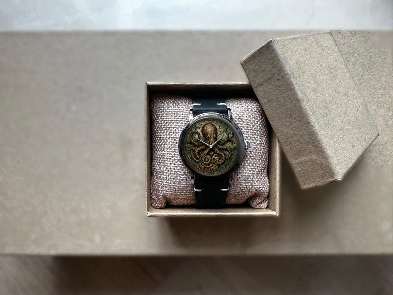 May include: A steampunk-style wristwatch with an octopus design on the face, presented in a small, open cardboard box. The watch has a black band and a detailed, antique-looking dial. The box is lined with a beige fabric.