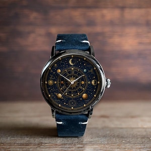 Könnte beinhalten: Eine runde Armbanduhr mit einem dunkelblauen Zifferblatt mit einem Himmelsdesign mit Planeten, Sternen und einem Sonnenstrahl. Die Uhr hat ein schwarzes Gehäuse, goldene Akzente und ein blaues Lederarmband. Die Uhr liegt auf einer Holzoberfläche.