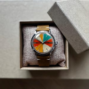 Op de afbeelding: Een vintage horloge met een kleurrijke, gesegmenteerde wijzerplaat in rood, geel, groen en wit. Het horloge heeft een zilveren kast en een bruine leren band. Het ligt in een kleine, open kartonnen doos op een neutrale ondergrond.