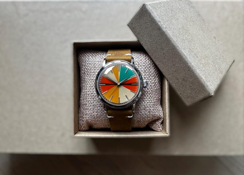 Op de afbeelding: Een vintage horloge met een kleurrijke, gesegmenteerde wijzerplaat in rood, geel, groen en wit. Het horloge heeft een zilveren kast en een bruine leren band. Het ligt in een kleine, open kartonnen doos op een neutrale ondergrond.