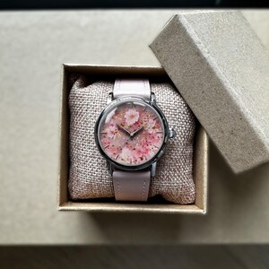 May include: A wristwatch with a pale pink leather strap and a round face displaying a floral pattern of pink blossoms and gold flecks. The watch is in an open cardboard box, on a burlap cushion.