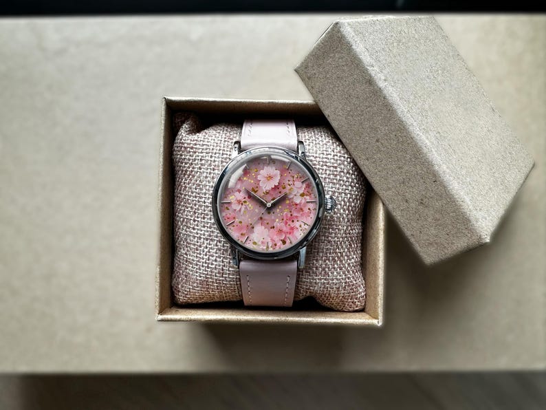 May include: A wristwatch with a pale pink leather strap and a round face displaying a floral pattern of pink blossoms and gold flecks. The watch is in an open cardboard box, on a burlap cushion.