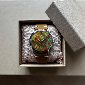 May include: A wristwatch with a colorful fish design on the face, set in a silver case with a brown leather band. The watch is presented in a small, open cardboard box on a neutral surface.