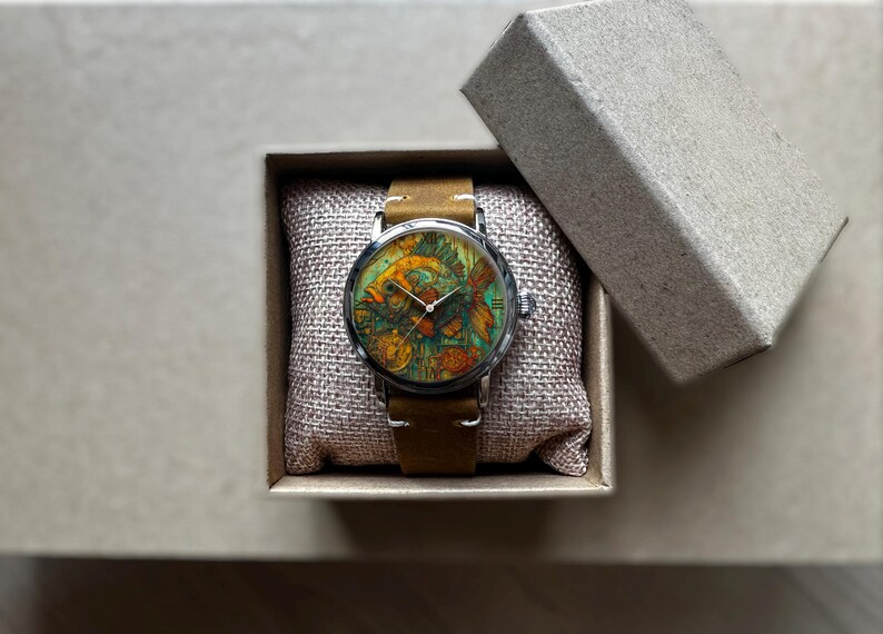 May include: A wristwatch with a colorful fish design on the face, set in a silver case with a brown leather band. The watch is presented in a small, open cardboard box on a neutral surface.