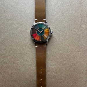 May include: A wristwatch with a brown leather band and a silver-colored case. The watch face features a colorful, segmented design with hues of red, orange, yellow, green, and blue. The band measures approximately 9.5 inches in length.