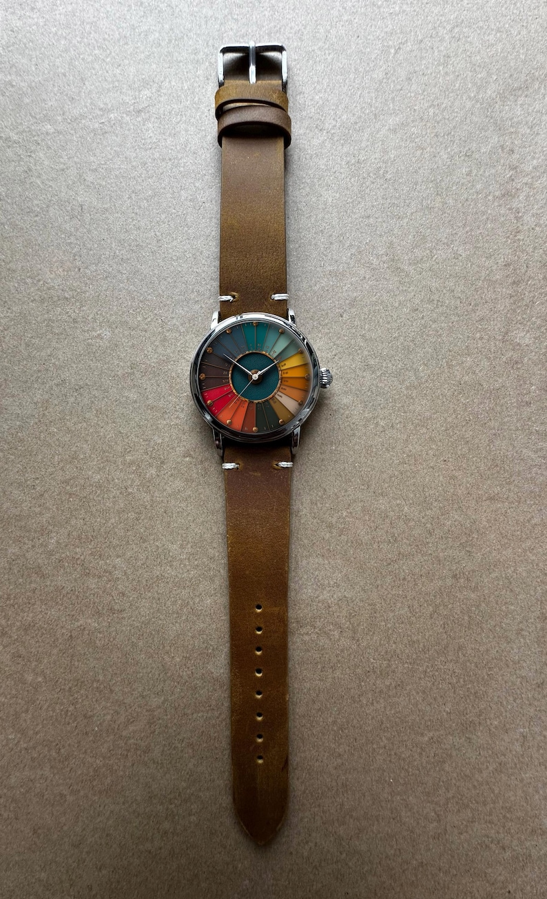 May include: A wristwatch with a brown leather band and a silver-colored case. The watch face features a colorful, segmented design with hues of red, orange, yellow, green, and blue. The band measures approximately 9.5 inches in length.