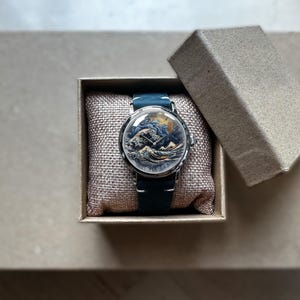 May include: A round wristwatch with a blue leather band and a detailed face depicting a seascape scene. The watch is presented in a small, open cardboard box with a textured, beige pillow.