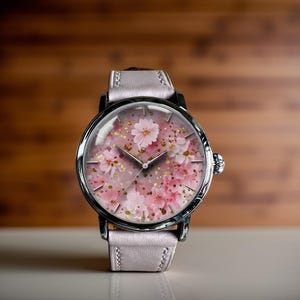 May include: A wristwatch with a pale pink leather strap and a silver case. The watch face displays a pink floral design with gold flecks. The watch hands are silver.