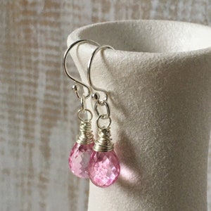 May include: Dangle earrings featuring faceted pink gemstones in a teardrop shape. The earrings have silver-coloured ear wires and decorative wire wrapping. The gemstones are displayed against a neutral background. The earrings are a delicate piece of jewellery.