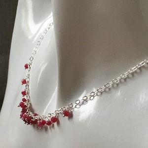 May include: A silver necklace featuring a fine chain and a collection of small, round, red beads. The beads are clustered, producing a cascading appearance. The necklace is shown on a white surface.