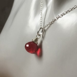May include: A silver necklace with a faceted, teardrop-shaped red gemstone pendant. The pendant is wire-wrapped and hangs from a delicate chain. The gemstone reflects light, highlighting its cut and colour.