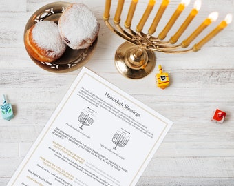 Könnte beinhalten: Eine goldene Menora mit brennenden gelben Kerzen, zwei mit Puderzucker bestäubte Krapfen in einer Metallschale, zwei Dreidels und eine gedruckte Anleitung für Hanukkah-Segenssprüche mit Diagrammen zum Anzünden der Kerzen auf einer weißen Holzoberfläche.