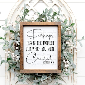 May include: A wooden sign with a white background and black text that reads "Perhaps this is the moment for which you were created. Esther 4:14". The sign is framed in a dark wood frame and is surrounded by a wreath of green leaves and white berries.
