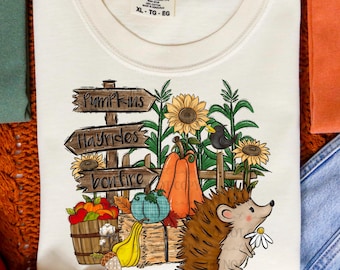 May include: Cream-colored Comfort Colors t-shirt with a fall-themed graphic. The design includes pumpkins, sunflowers, a hedgehog, and wooden signs that read "Pumpkins," "Hayrides," and "Bonfire."