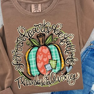 May include: A brown Comfort Colors sweatshirt with a pumpkin graphic. The pumpkin has a plaid section, hearts, and patches. The text reads "Your Hearts be Full" and "Thanks & Giving".
