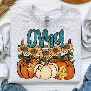 May include: A light gray sweatshirt with the word "OMA" in teal letters with white polka dots. The design includes sunflowers, pumpkins, and a wooden fence, perfect for fall. The sweatshirt is a Gildan brand.