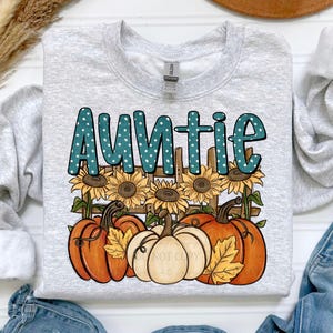 May include: Light grey sweatshirt with a fall design. The word "Auntie" is in teal polka-dot letters above sunflowers, pumpkins, and a wooden fence. The pumpkins are orange and white, creating a seasonal theme.
