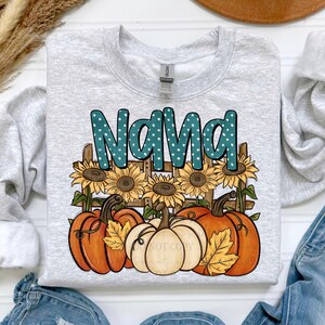 May include: A light gray sweatshirt with the word "NANA" in teal, polka-dotted letters. The graphic features sunflowers, pumpkins, and a wooden fence. The pumpkins are orange and white, with yellow leaves.