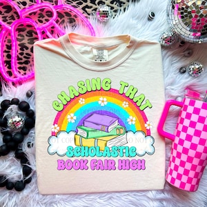 May include: Cream-colored t-shirt with a graphic that reads "Chasing That Scholastic Book Fair High." The design features a rainbow, books, and flowers. A pink checkered tumbler with a handle is on the right.