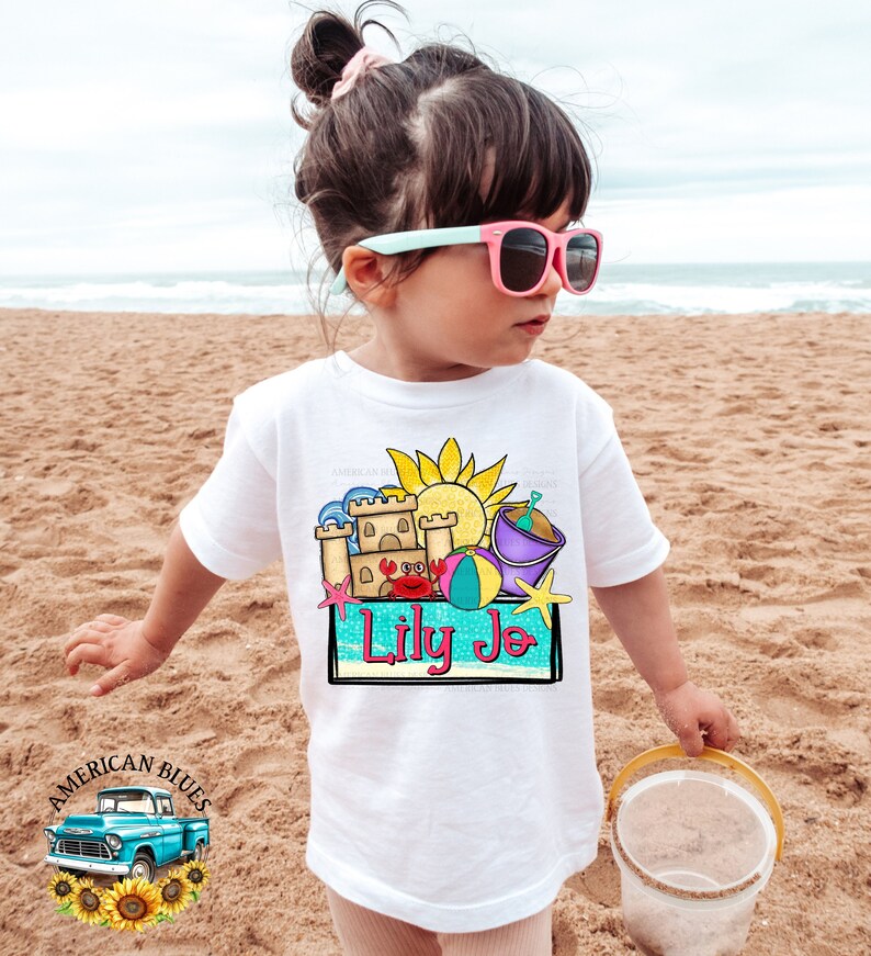 May include: White t-shirt with a colorful graphic of a sandcastle, beach ball, sun, and pail. The text "Lily Jo" is written in a blue box on the graphic.