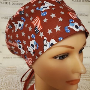 Könnte beinhalten: Rote chirurgische Mütze mit einem Muster aus weißen Hunden, die blaue und rote amerikanische Flaggen-Bandanas und silberne Sterne tragen. Die Mütze hat eine rote Schleife.