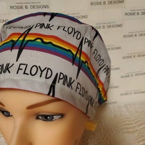 Op de afbeelding: Een witte chirurgische scrub-muts met een regenboogstreep en de tekst "Pink Floyd" herhaald in zwart.