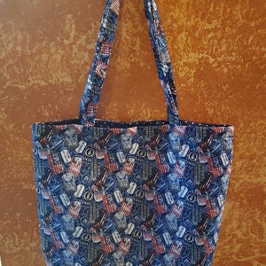 Può includere: Una borsa tote blu con un motivo di bandiere americane e targhette militari. La borsa ha due lunghi lacci.